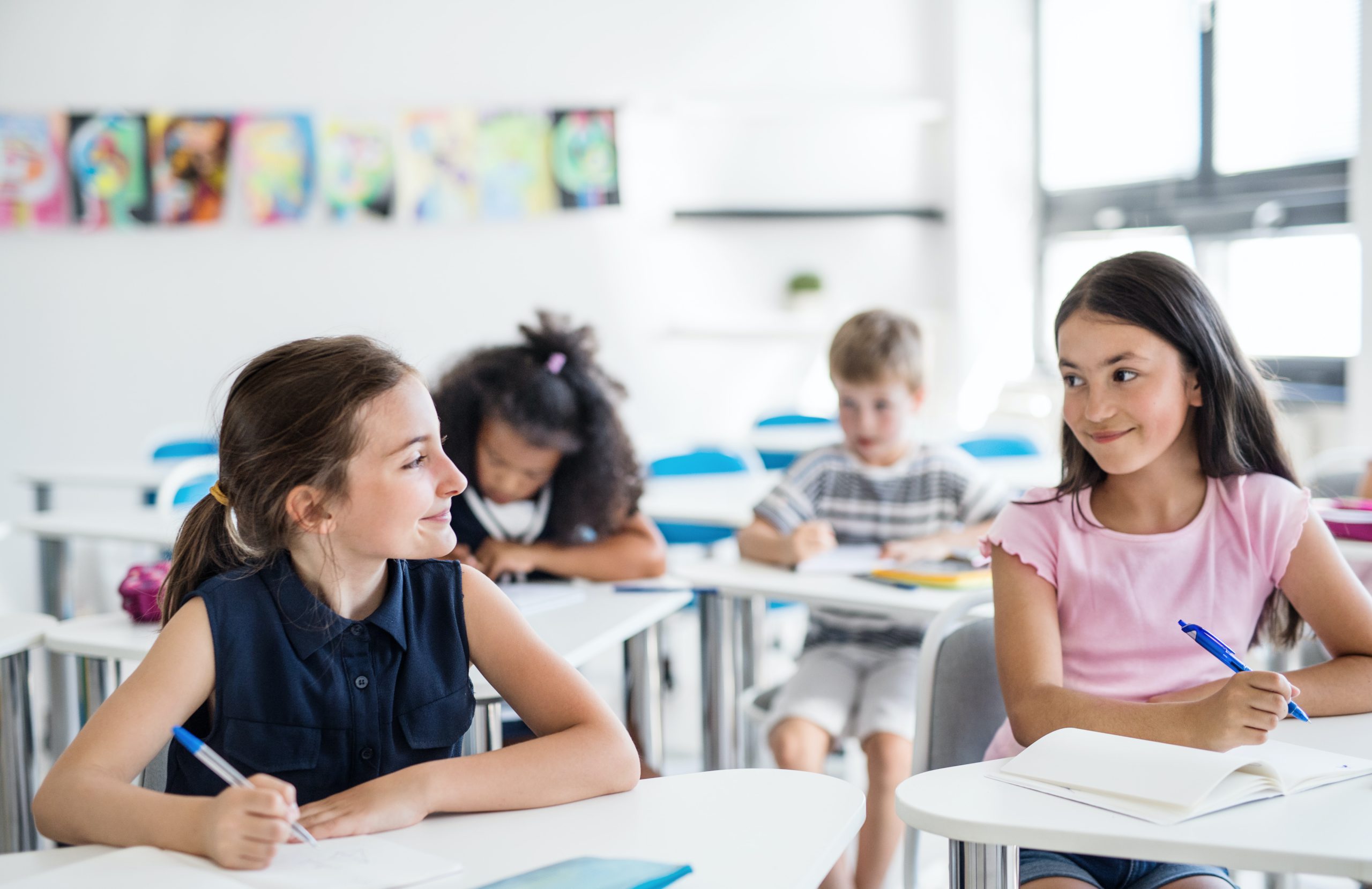 Two young girls smile at each other as they sit at their desks in a classroom, ready to pray a one-minute prayer for a new school year.
