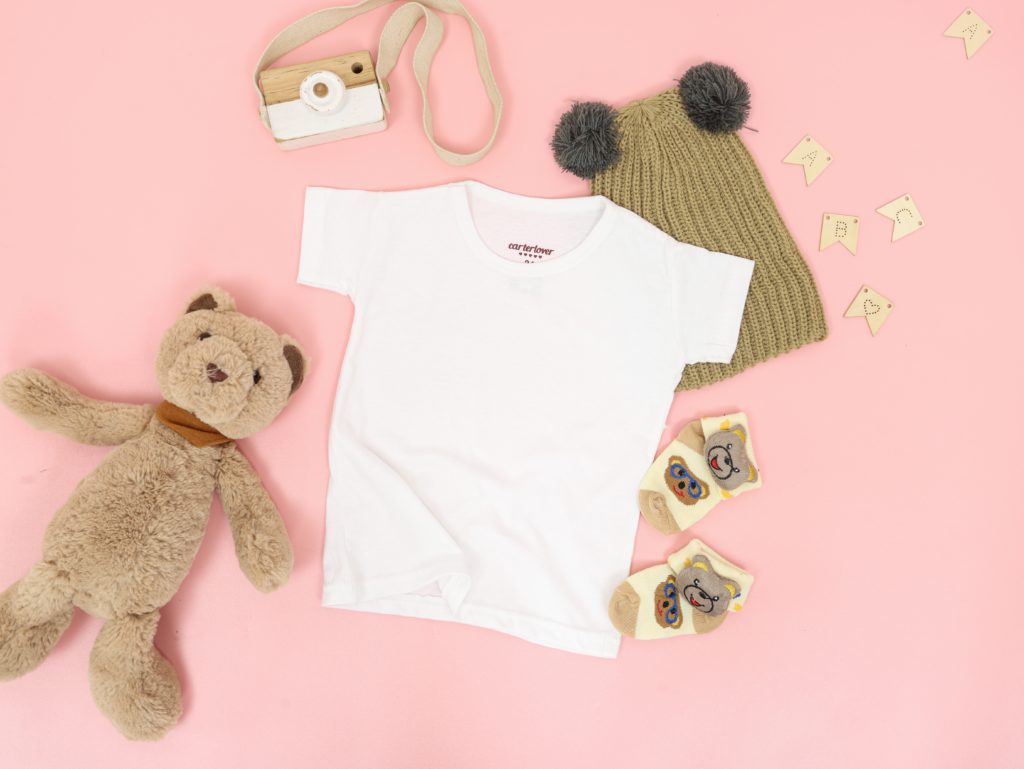 a white baby T-shirt with stuff bear, hat and socks sit on a pink background reminding you to pray a one-minute prayer for a new baby.