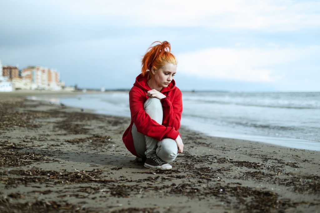 A young woman wearing a red sweatshirt sits on the beach praying a short prayer for social anxiety.