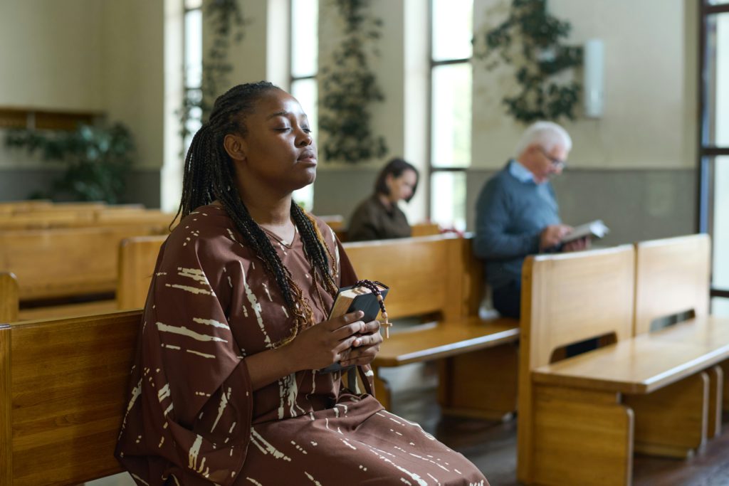 A Black woman with her eyes closed holds a Bible in a church pew with a posture of humility.