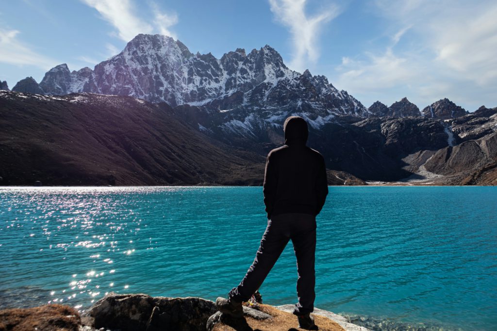 A man facing snowing peaks across a blue sea stands in awe of God.