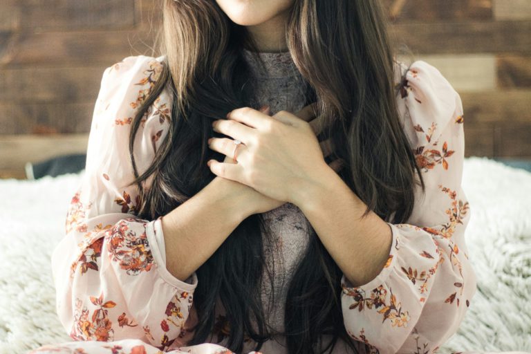 A young woman with long dark hair in s a flowered shirt holds her hands over her chest showing how to surrender your heart to God.