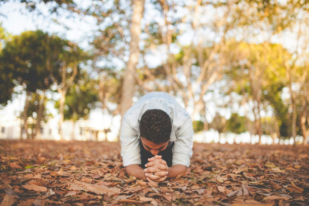 A man in a white shirt kneels with his forearms in the dirt and his hands clasped in prayer as he is battling doubts.