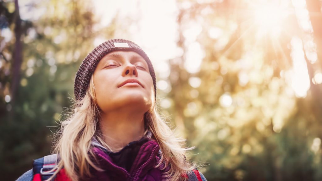 A young woman who has learned to choose contentment wherever she is closes her eyes and breathes in deeply while outside in the sunshine in the woods.