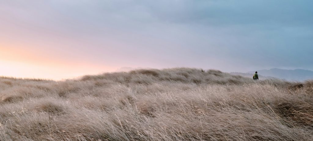 I man walking at sunrise on a brown hill of grass is finding contentment in God.