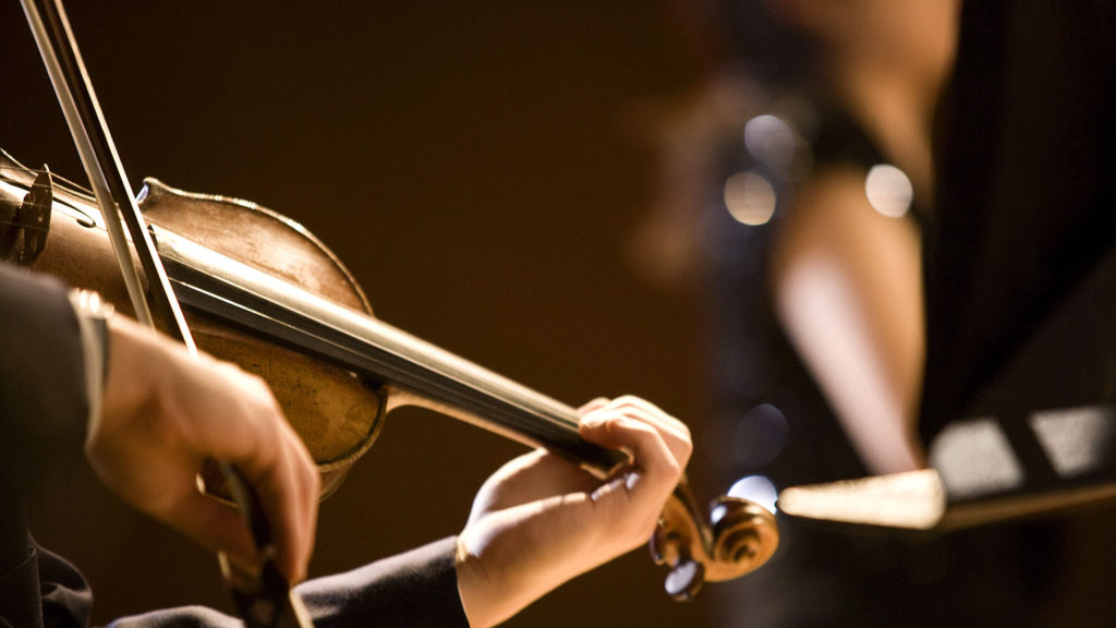 A sepia-tone image of hands playing a violin in harmony with an orchestra.