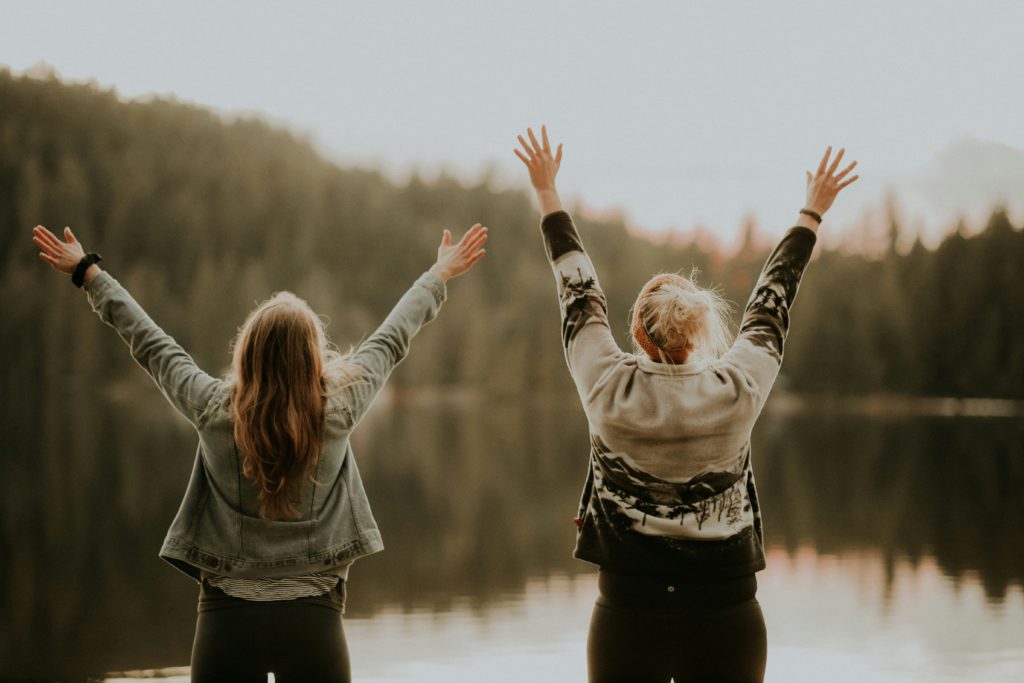 Two women stand before a mountain lake at sunset with raised arms as they realize they are more than enough.