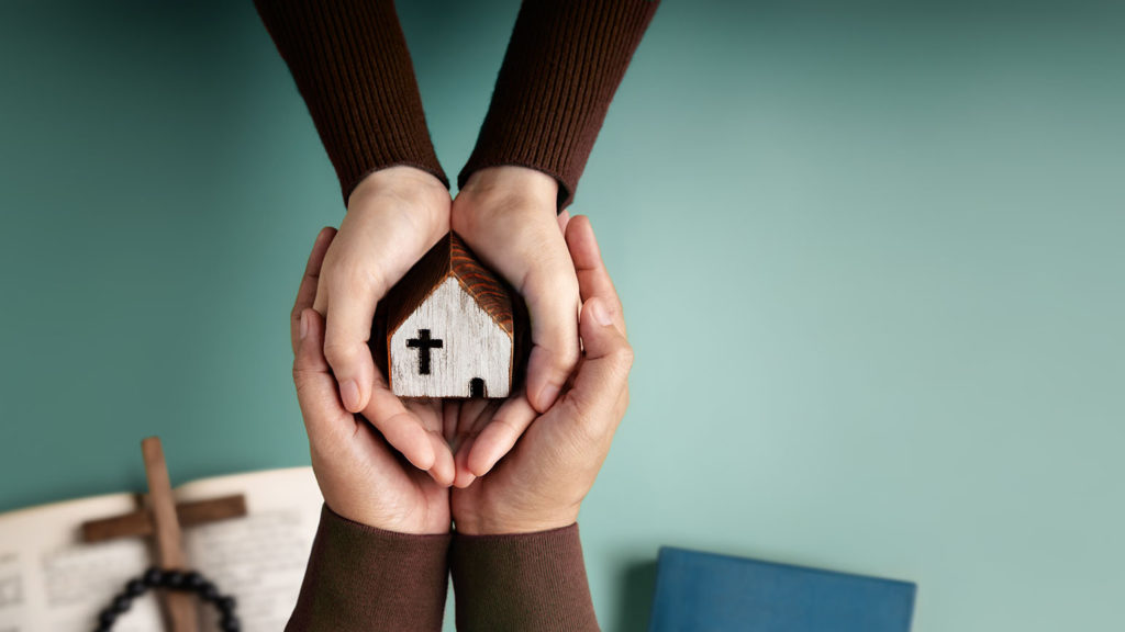 Two pairs of hands cup a small wooden house shared church where we can show love in action.