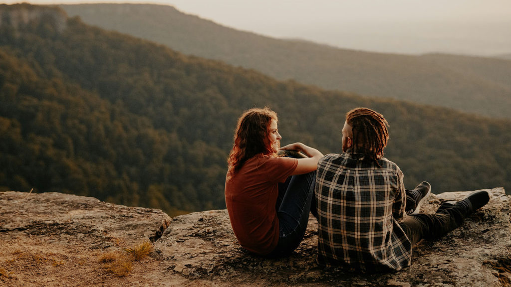 A woman and man sit on a mountain ledge together as they show kindness to one another.