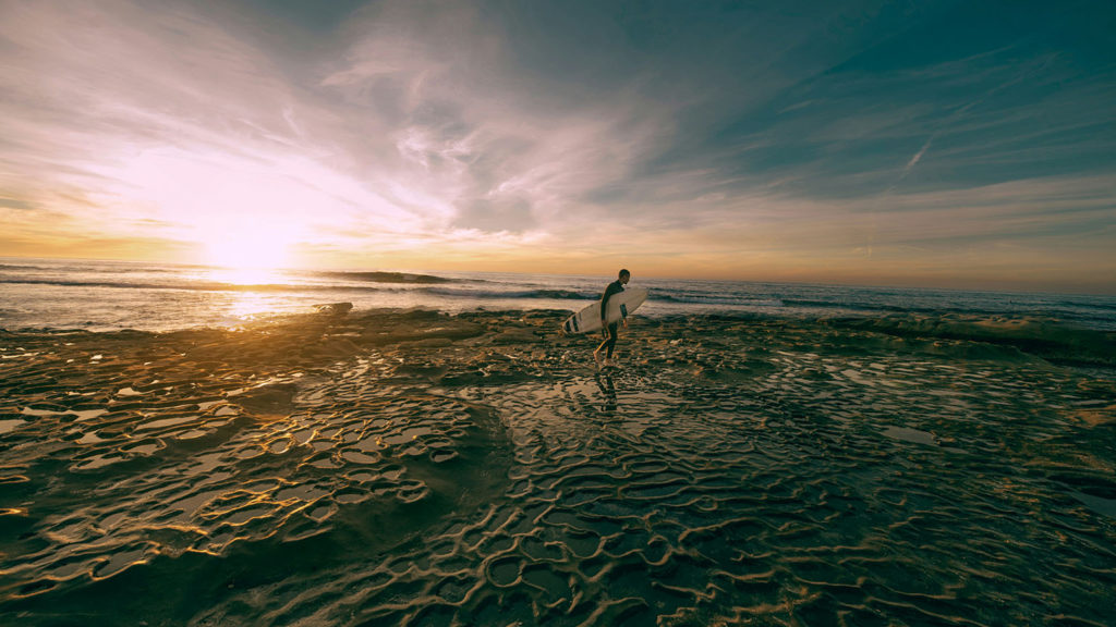 A young man carries his surfboard on the beach at sunrise because he got good sleep and energy abounds.