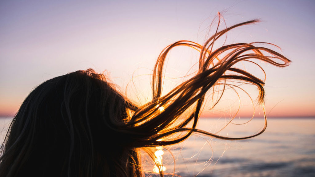 A woman's hair flies in the wind as she stands at the ocean at sunset knowing the she is loved by God spirit, soul, and body.